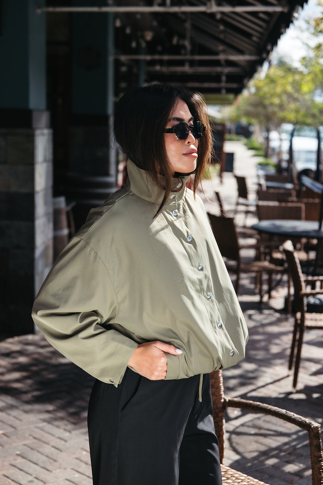 Woman wearing the Naia Funnel Neck Jacket, a lightweight light olive snap-front jacket with a drawstring hem