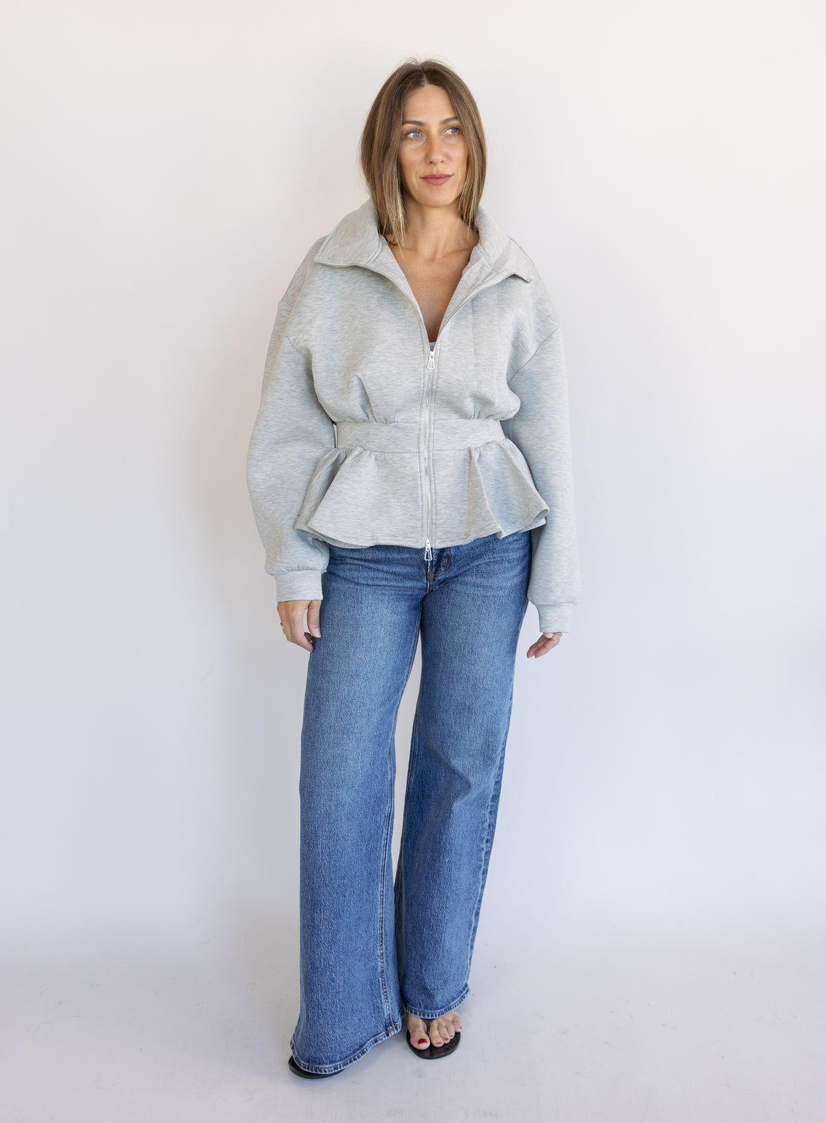 Woman wearing the Annika Peplum Zip Jacket, a heather grey knit zip-up with a funnel collar and peplum hem