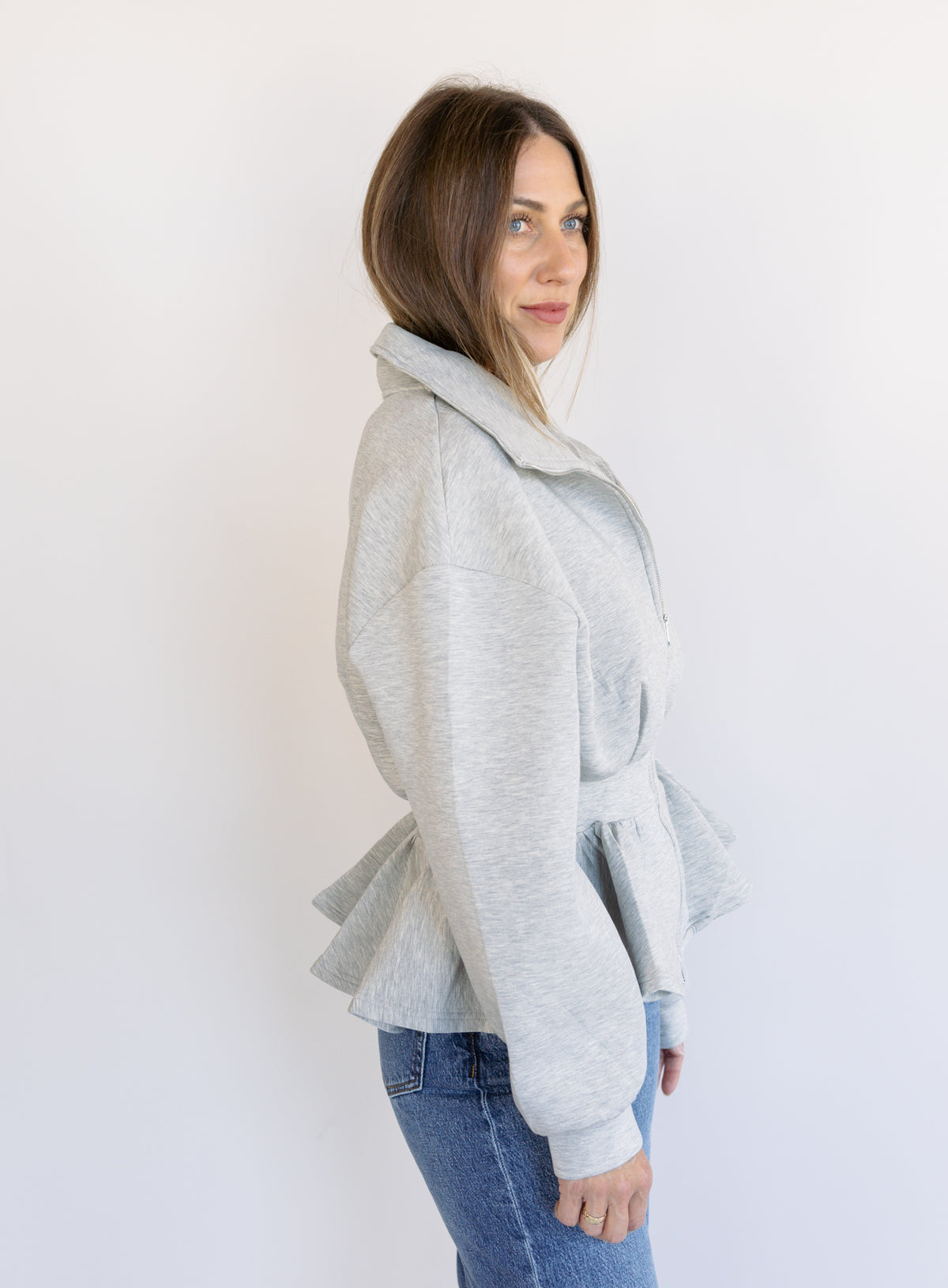 Woman wearing the Annika Peplum Zip Jacket, a heather grey knit zip-up with a funnel collar and peplum hem