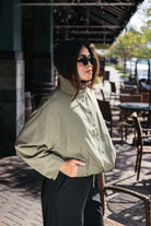 Woman wearing the Naia Funnel Neck Jacket, a lightweight light olive snap-front jacket with a drawstring hem