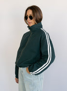 Woman wearing the Marina Twill Track Jacket, a forest green zip-up jacket with white sleeve stripes