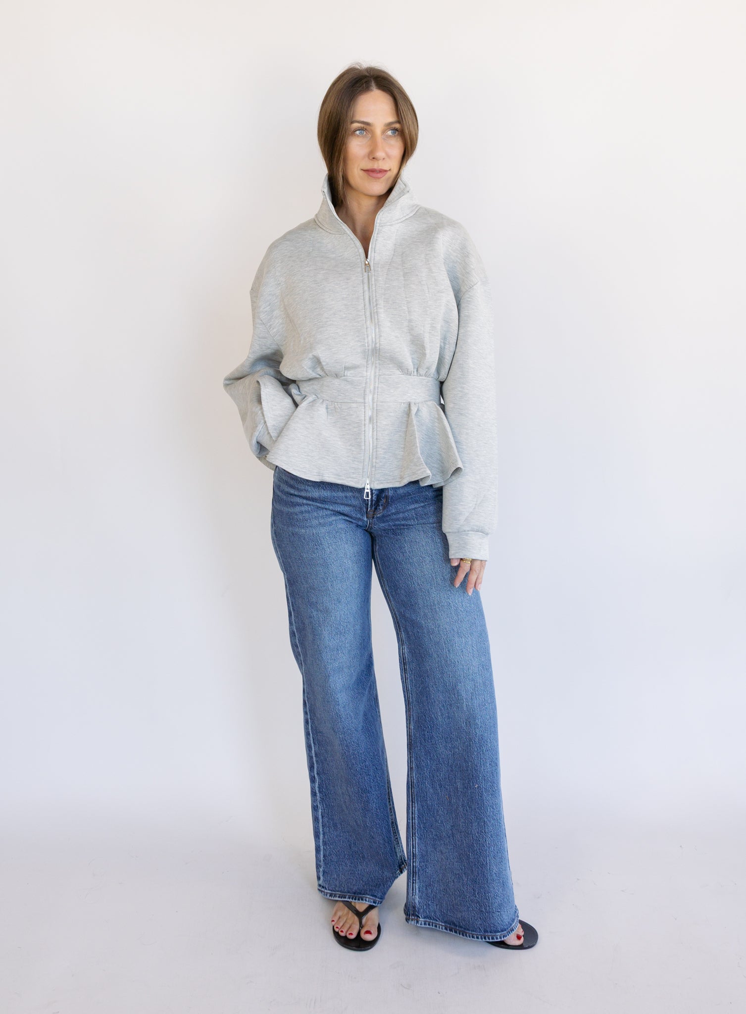 Woman wearing the Annika Peplum Zip Jacket, a heather grey knit zip-up with a funnel collar and peplum hem