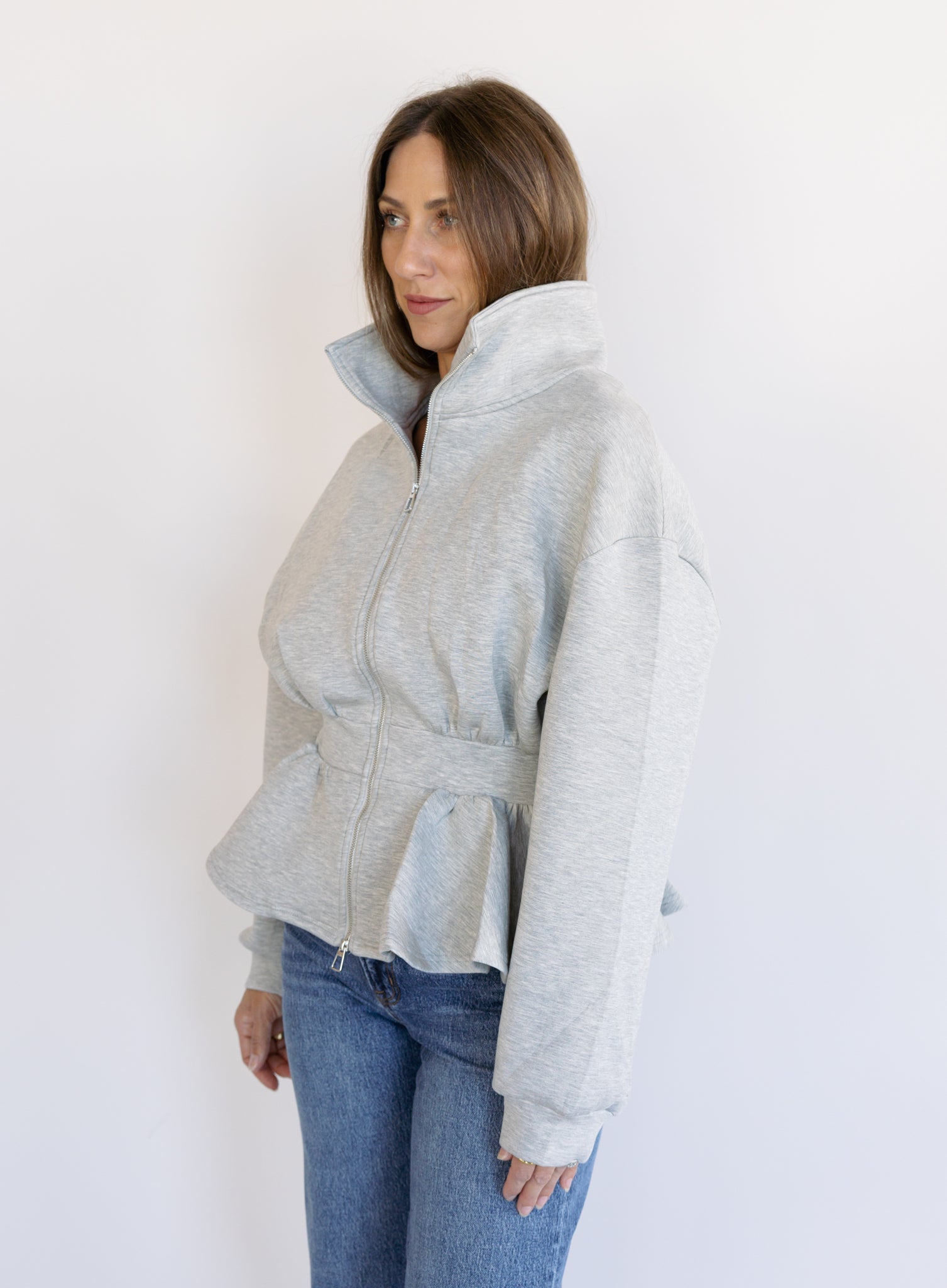 Woman wearing the Annika Peplum Zip Jacket, a heather grey knit zip-up with a funnel collar and peplum hem