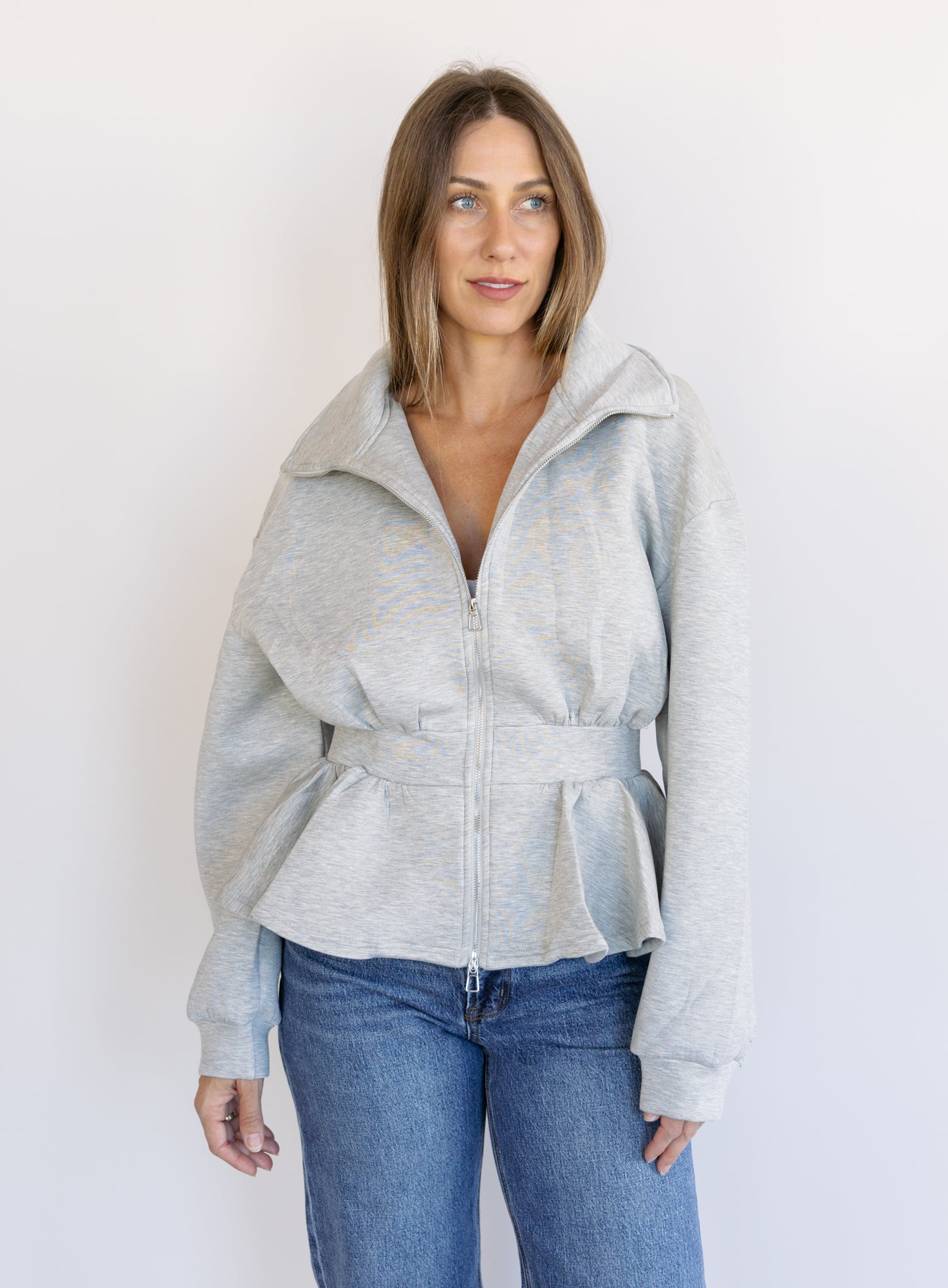 Woman wearing the Annika Peplum Zip Jacket, a heather grey knit zip-up with a funnel collar and peplum hem