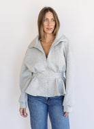 Woman wearing the Annika Peplum Zip Jacket, a heather grey knit zip-up with a funnel collar and peplum hem
