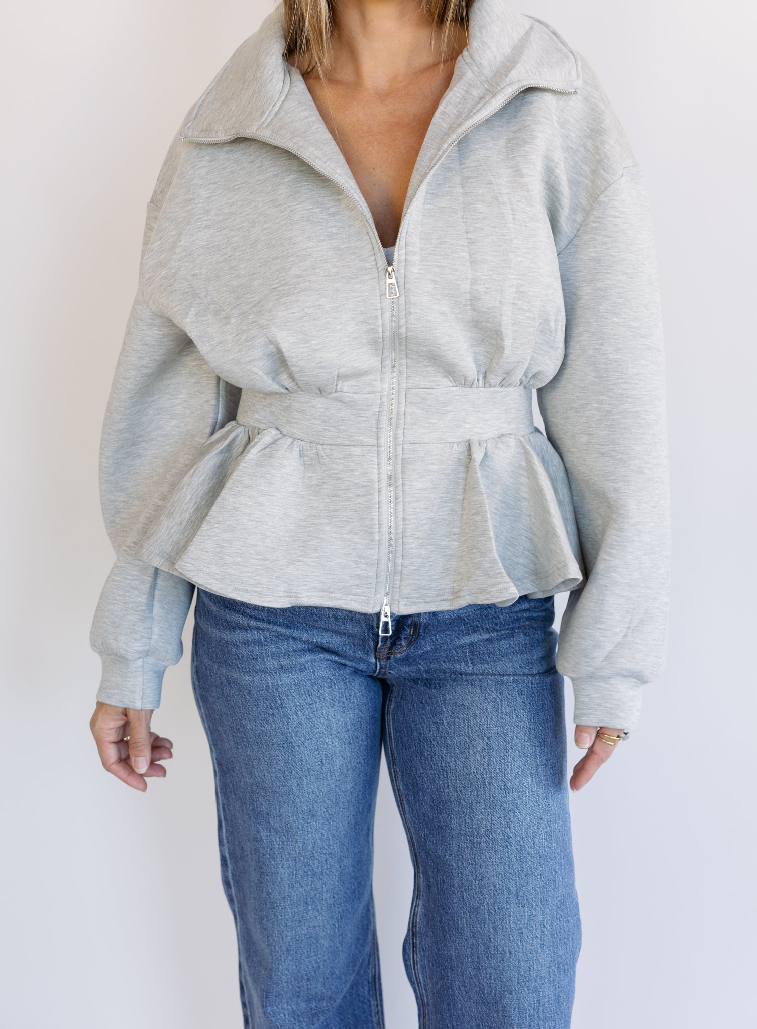 Woman wearing the Annika Peplum Zip Jacket, a heather grey knit zip-up with a funnel collar and peplum hem