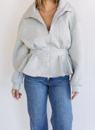 Woman wearing the Annika Peplum Zip Jacket, a heather grey knit zip-up with a funnel collar and peplum hem