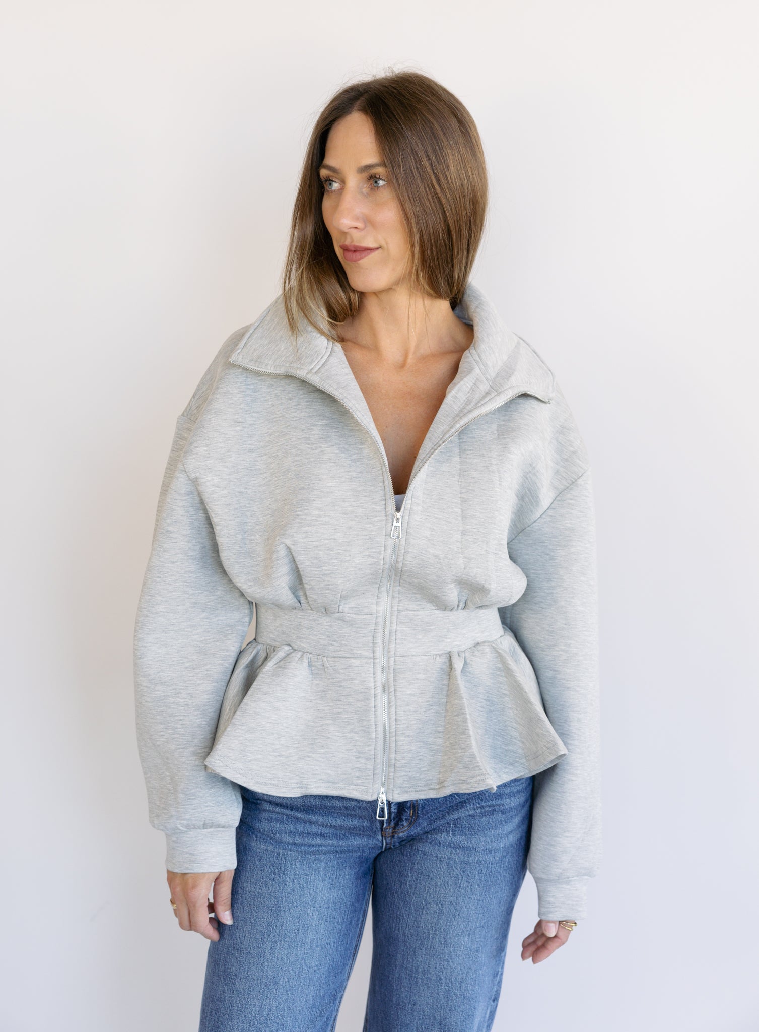 Woman wearing the Annika Peplum Zip Jacket, a heather grey knit zip-up with a funnel collar and peplum hem