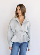 Woman wearing the Annika Peplum Zip Jacket, a heather grey knit zip-up with a funnel collar and peplum hem