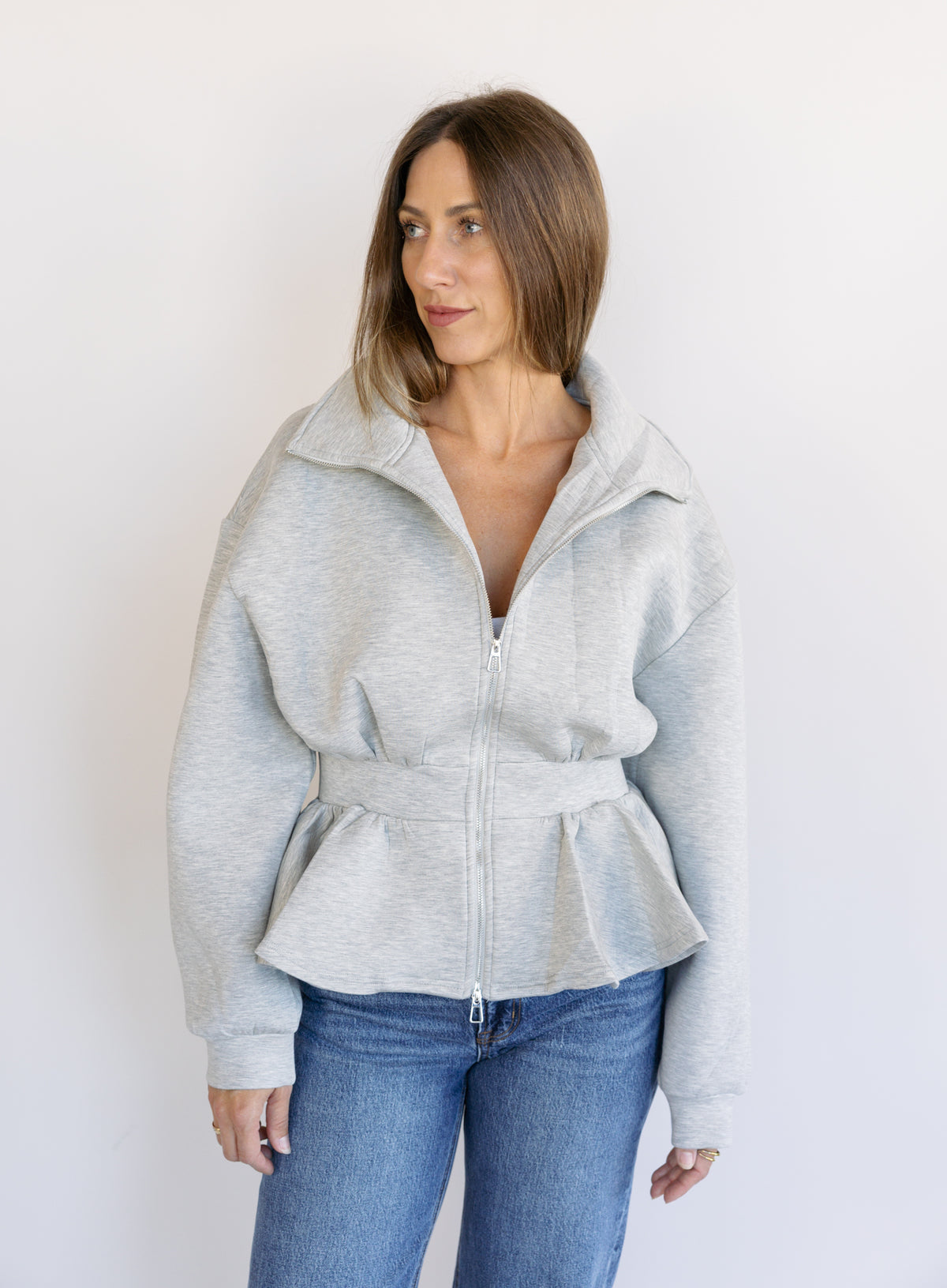 Woman wearing the Annika Peplum Zip Jacket, a heather grey knit zip-up with a funnel collar and peplum hem