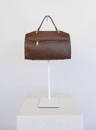 Beatrice Satchel, a brown ostrich-embossed top-handle bag with a gold bar clasp